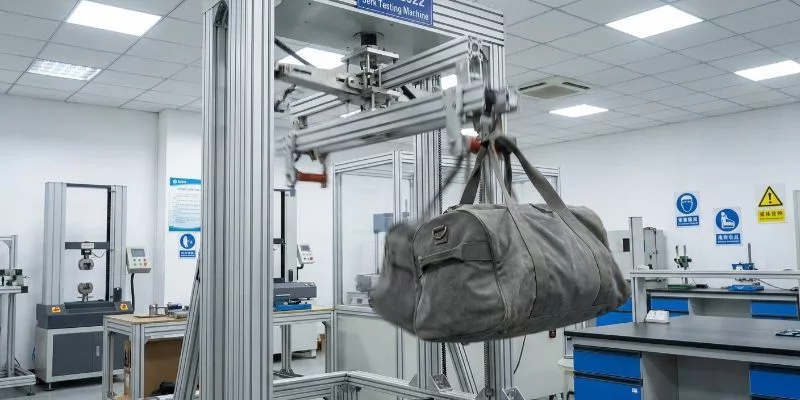 QB/T 2922 Jerk Testing Machine violently dropping and catching a loaded grey duffle bag in a QC lab.