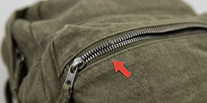 A red defect arrow sticker pointing to a broken metal zipper on a canvas backpack.