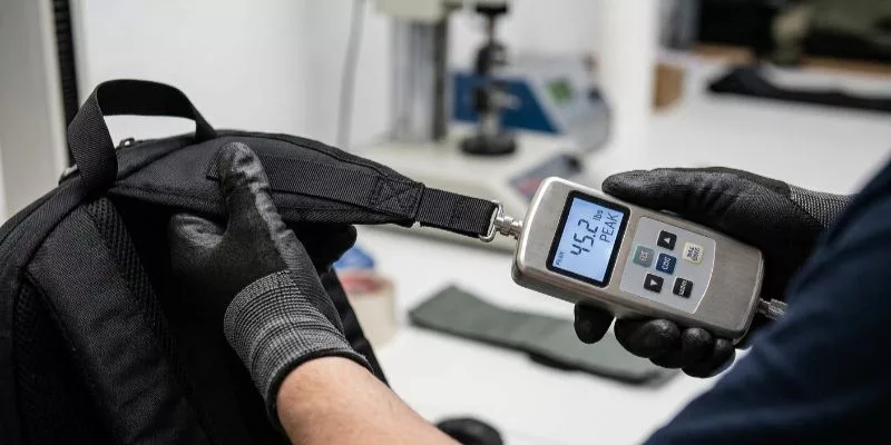 Inspector performing a destructive pull test on a backpack strap using a digital force gauge tool.